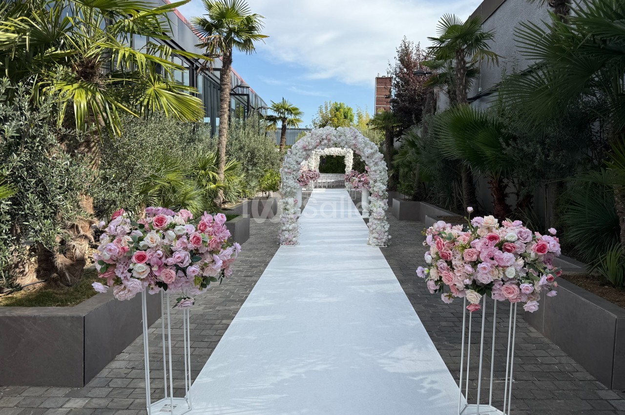 Allée extérieure décorée pour un mariage, avec arche florale blanche et arrangements de fleurs roses sur supports.