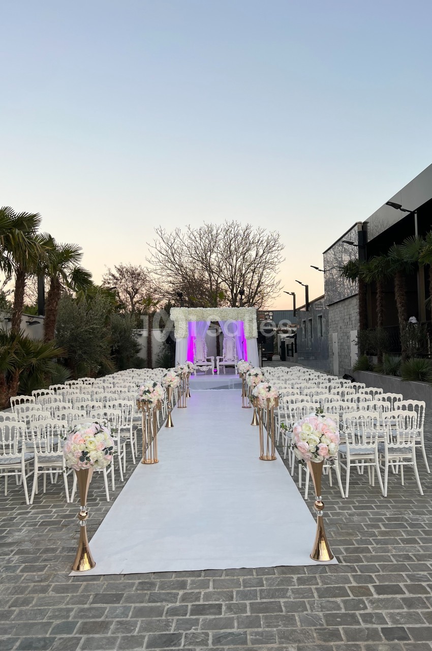 Allée centrale décorée de fleurs menant à une arche de mariage illuminée, entourée de chaises blanches en extérieur.