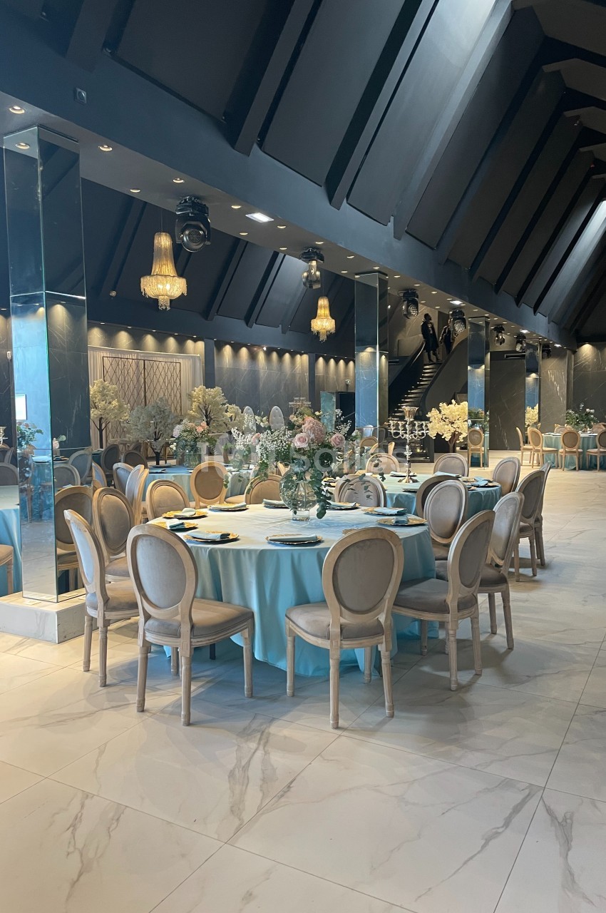 Salle de réception élégante avec tables rondes dressées, nappes bleues, chaises en bois clair et décorations florales.