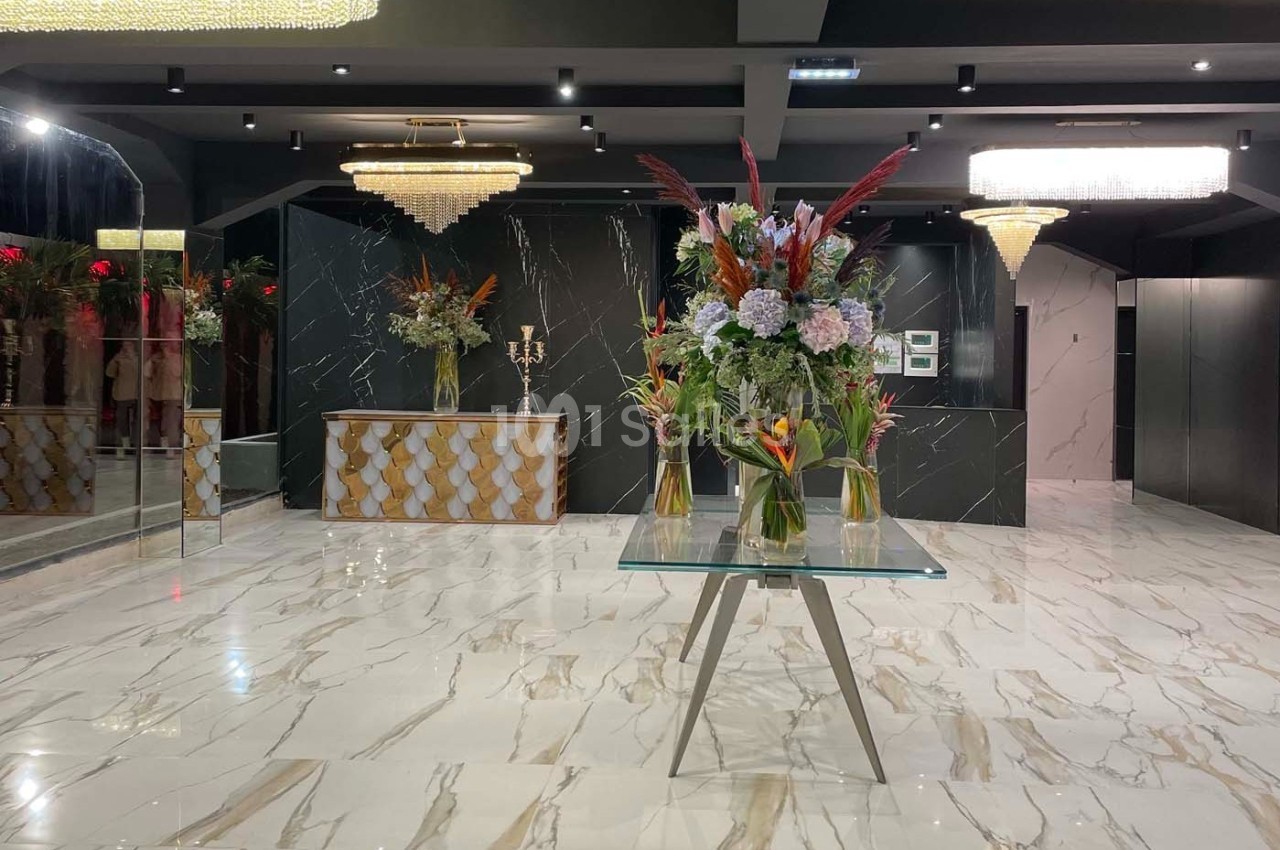 Hall élégant avec sol en marbre, comptoir décoratif, lustres et table centrale ornée de grands bouquets de fleurs.