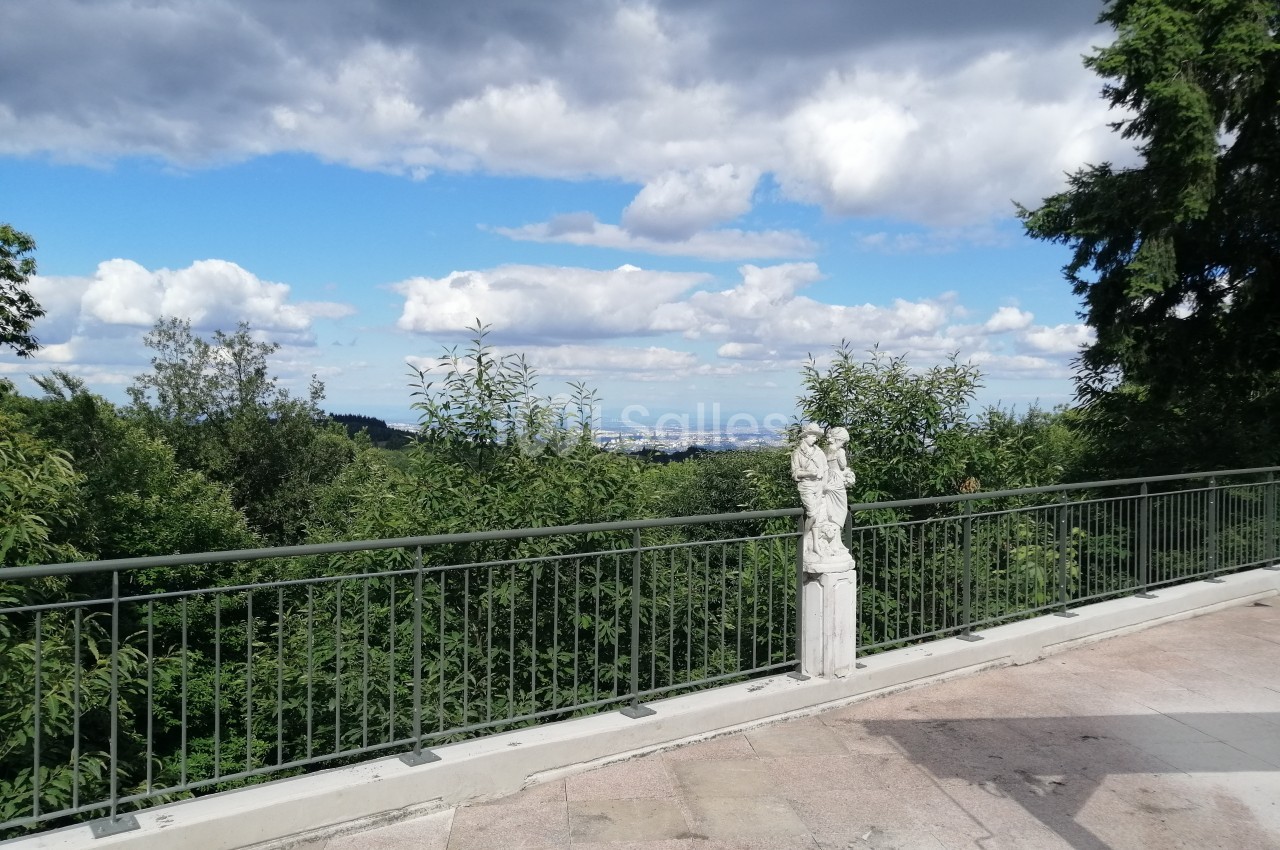 Vue d'une terrasse avec une statue, une balustrade, des arbres et un ciel partiellement nuageux en arrière-plan.