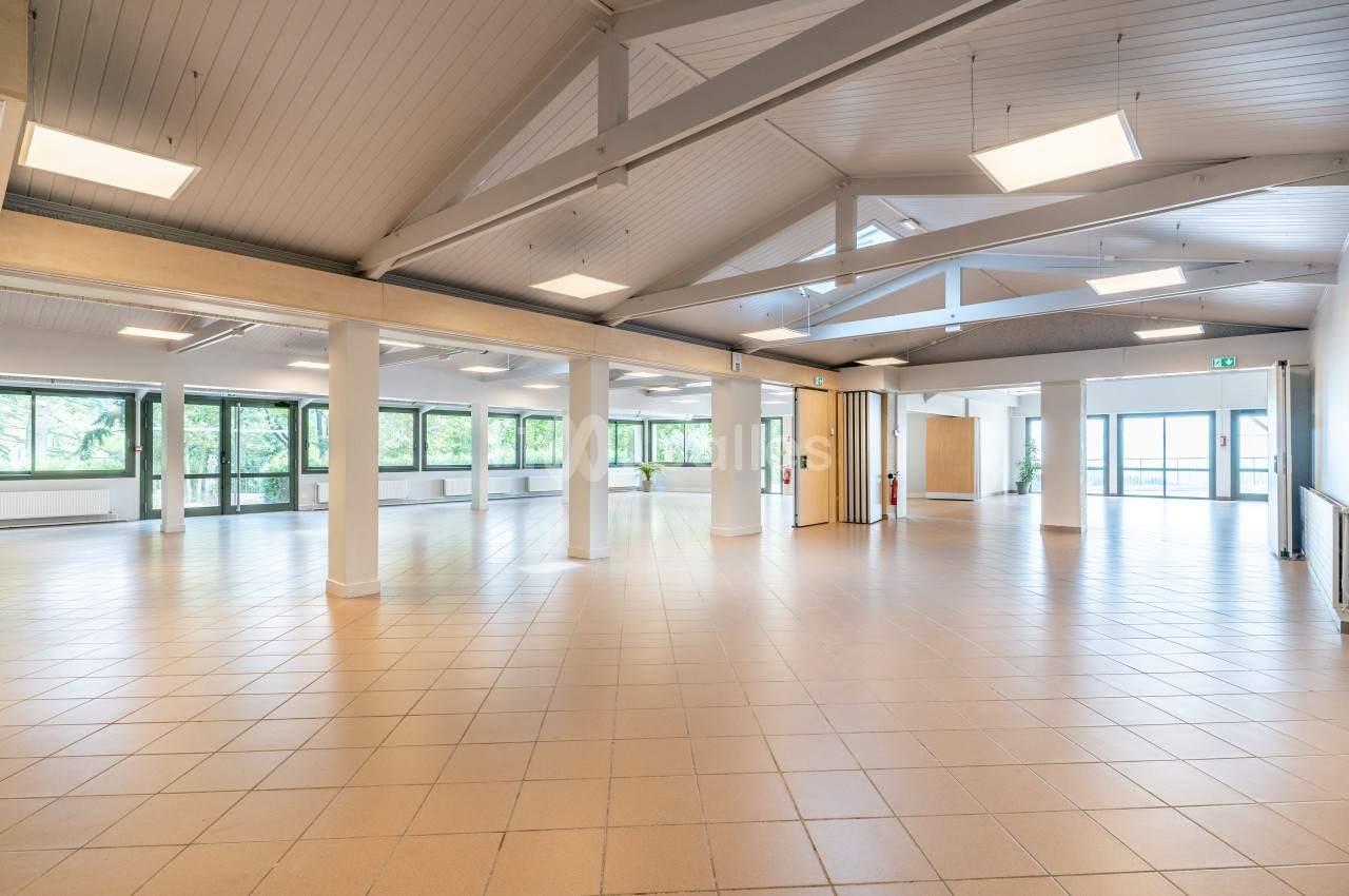Salle lumineuse avec plafond en poutres apparentes, carrelage beige et grandes fenêtres donnant sur un espace verdoyant.