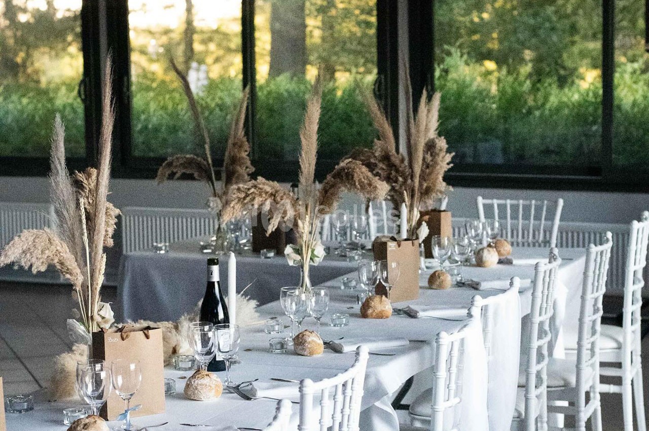 Tables décorées pour un événement, avec nappes blanches, pampas, bouteilles, verres et petits pains disposés.