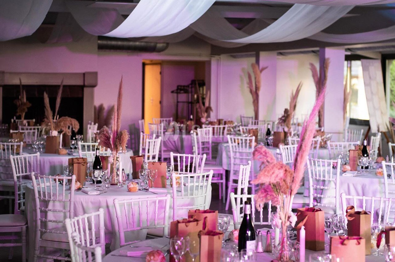 Salle de réception décorée avec des tables rondes, nappes blanches, chaises blanches et ornements floraux roses.