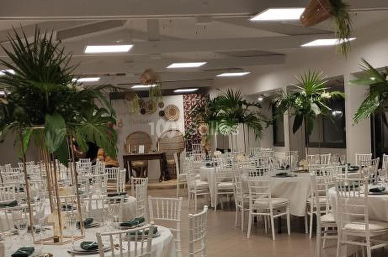 Salle de réception décorée avec des tables rondes, nappes blanches, chaises blanches et plantes tropicales.