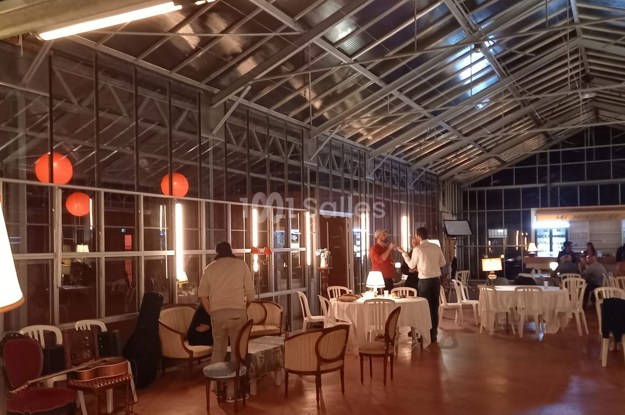 Salle vitrée avec tables et chaises, éclairée par des lampes et lanternes rouges, des personnes discutent.