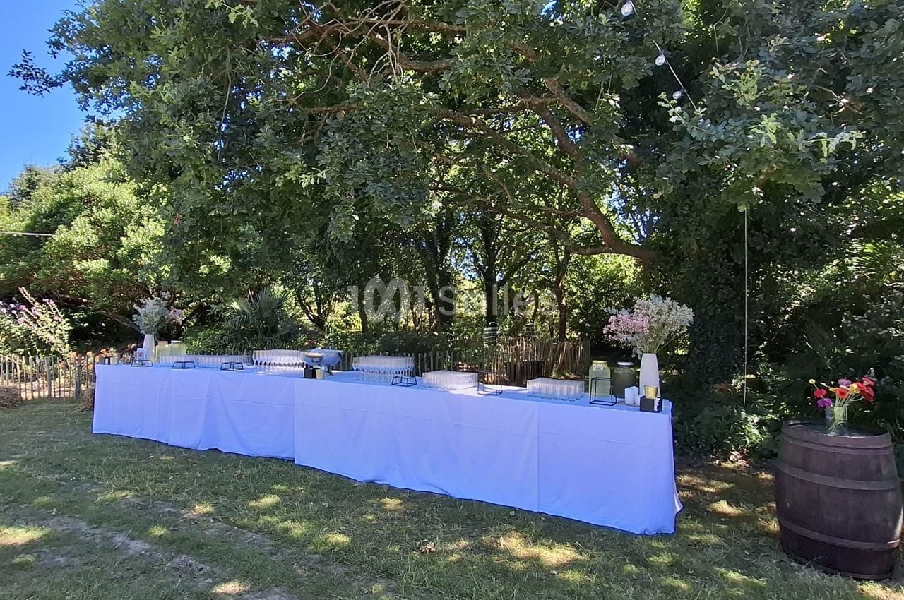 Table dressée avec nappes blanches en extérieur, sous des arbres, décorée de fleurs et entourée de verdure.