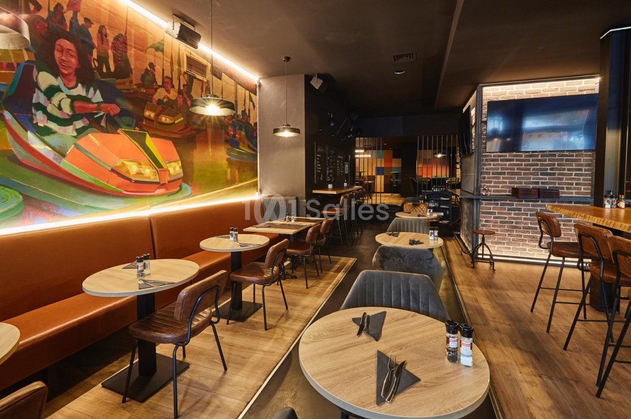 Salle de restaurant moderne avec tables en bois, chaises en cuir, fresque colorée et éclairage chaleureux.