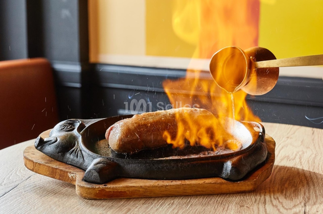Saucisse flambée dans une poêle en fonte, avec une sauce versée depuis une louche en métal.