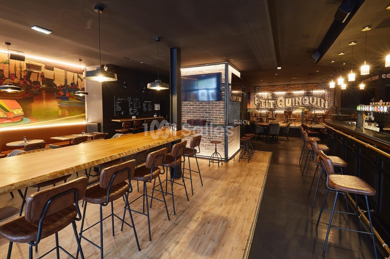 Intérieur d'un bar-restaurant moderne avec comptoir, tables en bois, chaises hautes et éclairage chaleureux.