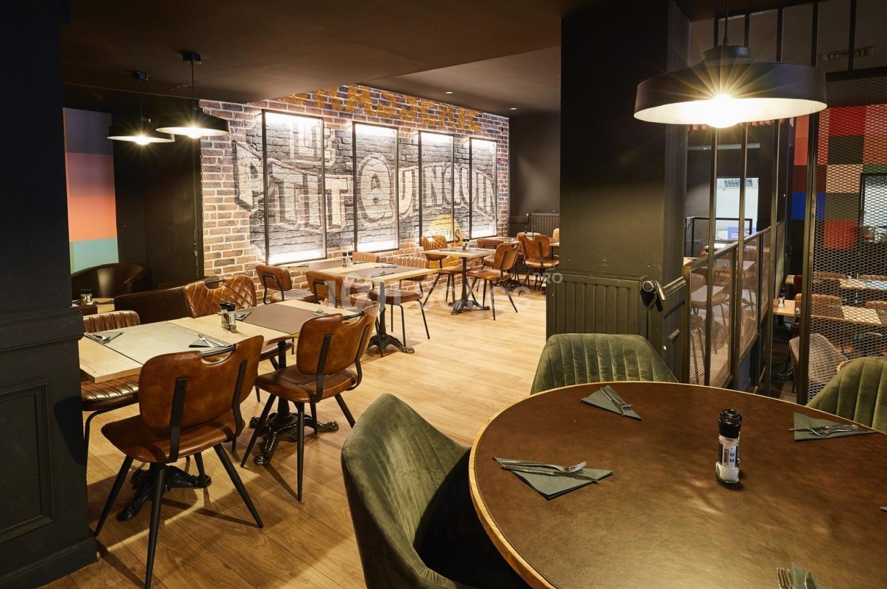 Salle de restaurant moderne avec murs en briques, tables en bois, chaises en cuir et éclairage tamisé.