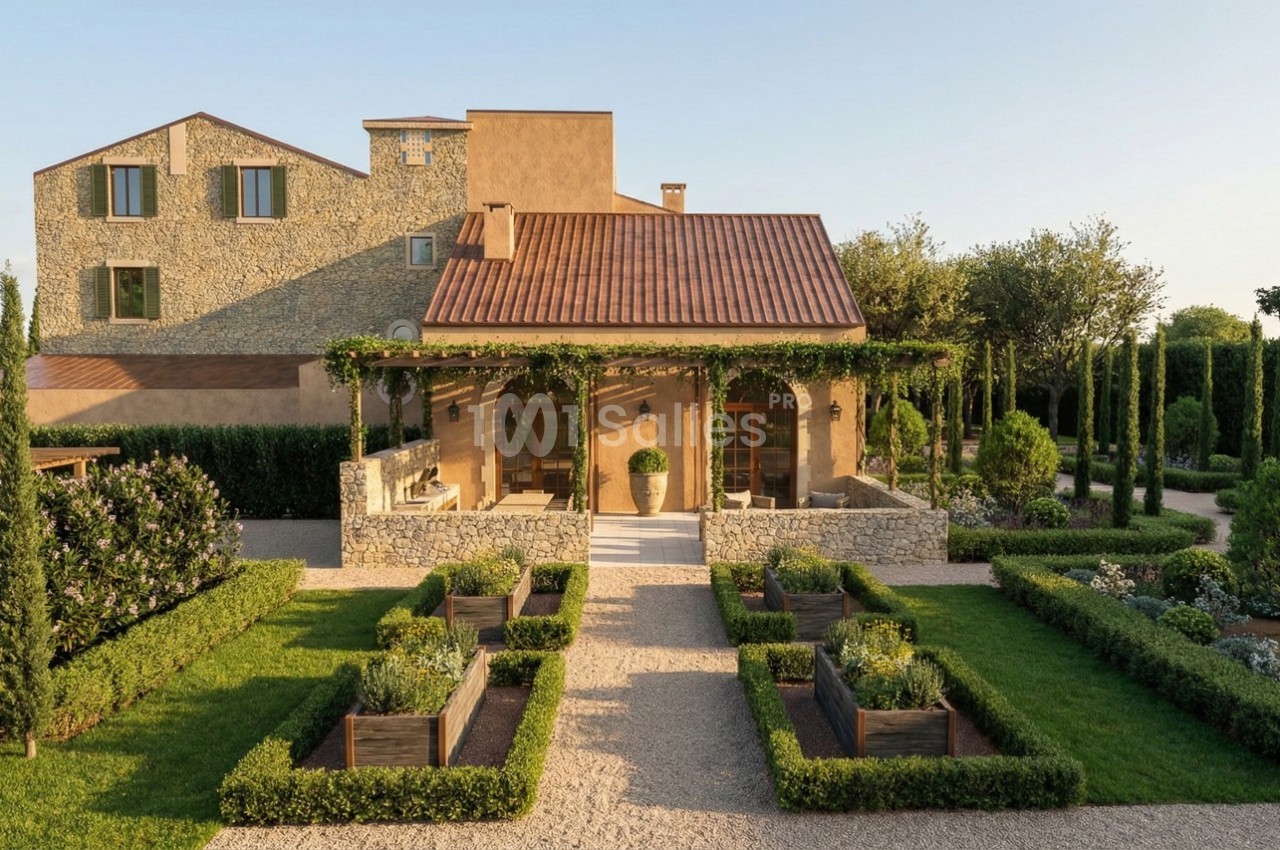 Façade d'une maison en pierre avec terrasse couverte, entourée d'un jardin aménagé et de chemins gravillonnés.