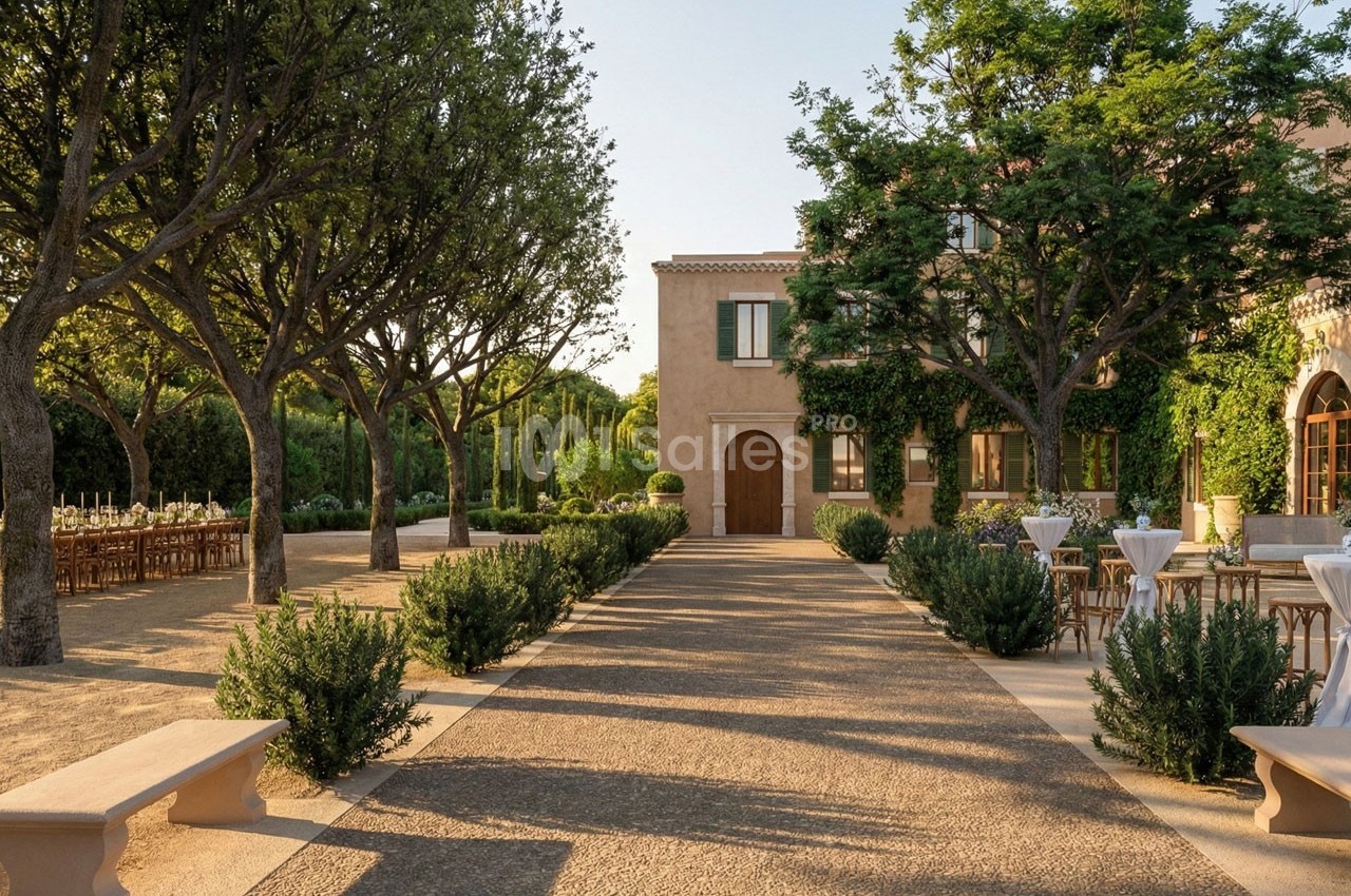 Allée bordée d'arbres menant à une maison en pierre, avec des tables dressées en extérieur sur les côtés.