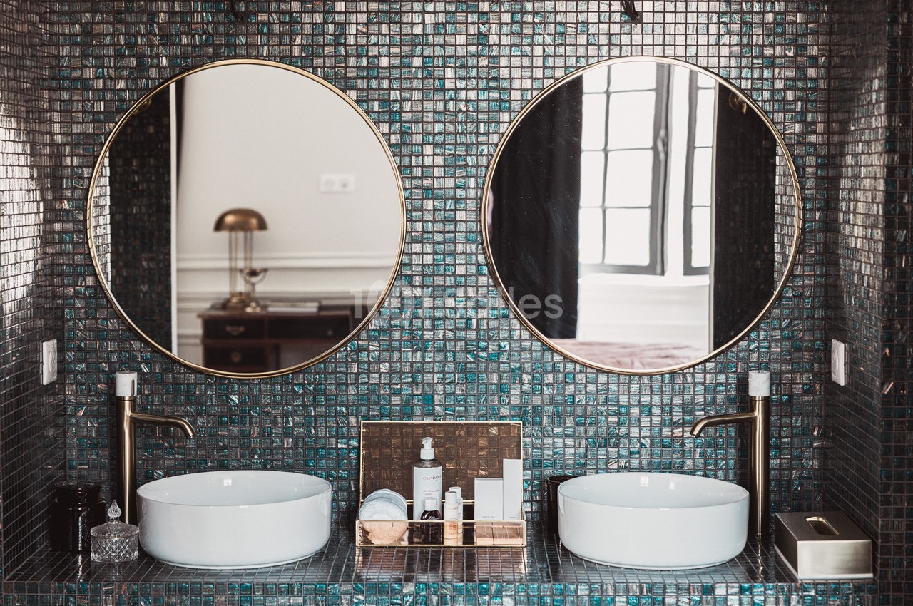 Deux lavabos blancs avec robinets dorés devant un mur en mosaïque sombre, surmontés de miroirs ronds.