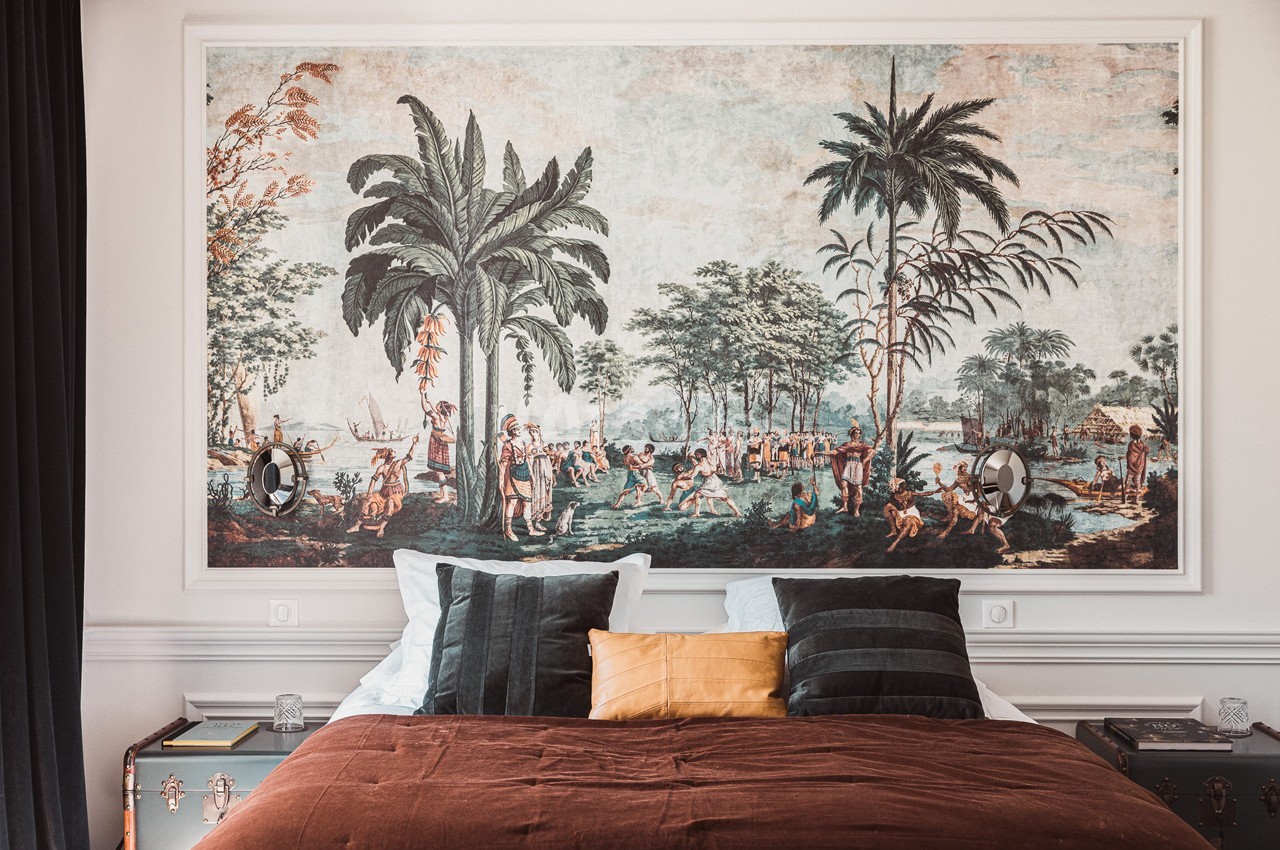 Lit avec coussins devant une grande fresque murale représentant une scène tropicale avec palmiers et personnages.