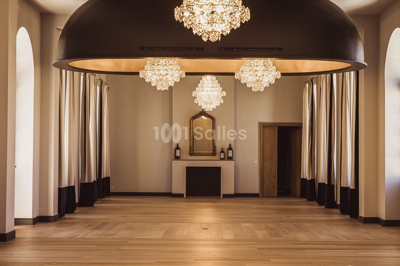 Salle élégante avec parquet en bois, lustres en cristal suspendus, rideaux encadrant des fenêtres et cheminée décorative.