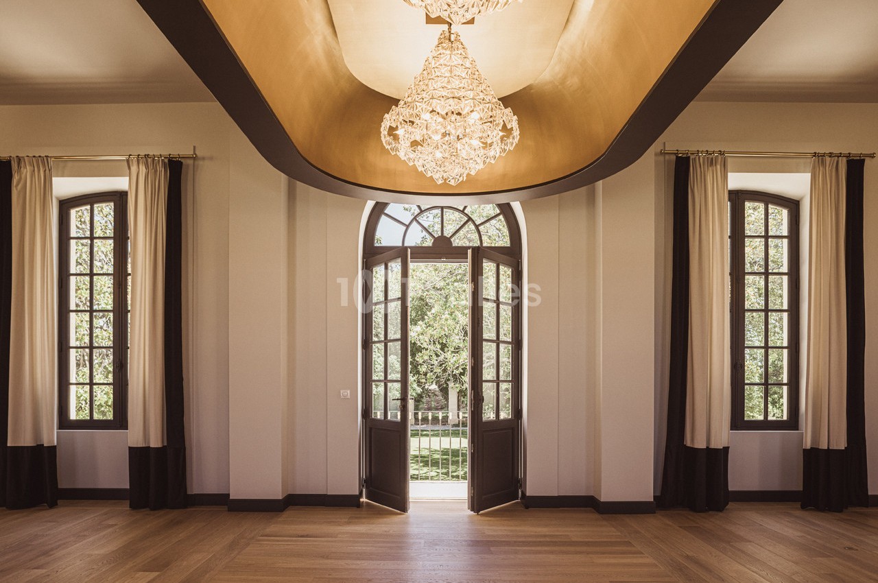 Salle lumineuse avec parquet, lustre central, grandes fenêtres et porte-fenêtre donnant sur un espace extérieur verdoyant.