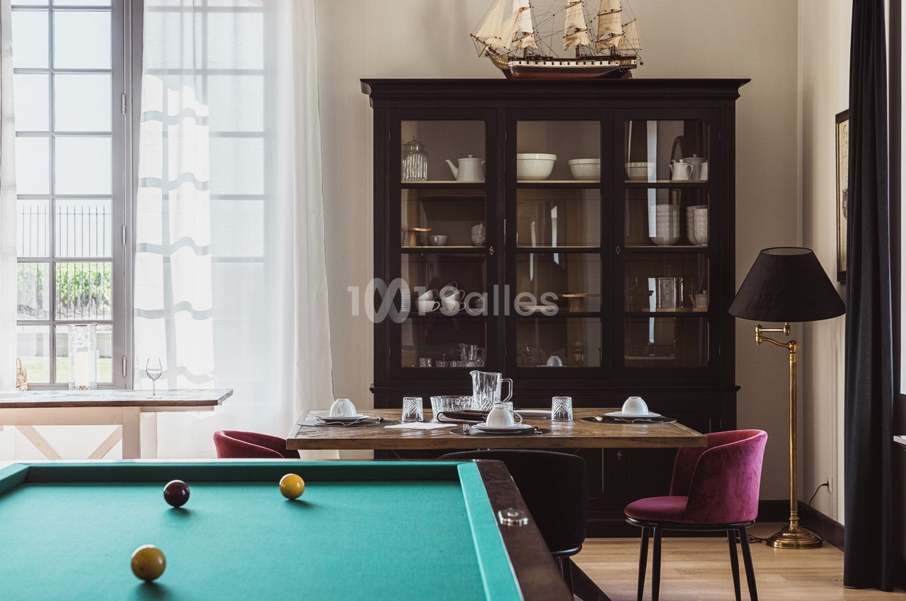 Salle à manger avec table dressée, buffet vitré, lampe sur pied et table de billard au premier plan.