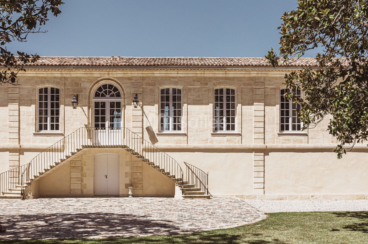 Façade d'un bâtiment en pierre avec escalier central, grandes fenêtres et cour pavée, entouré de verdure.