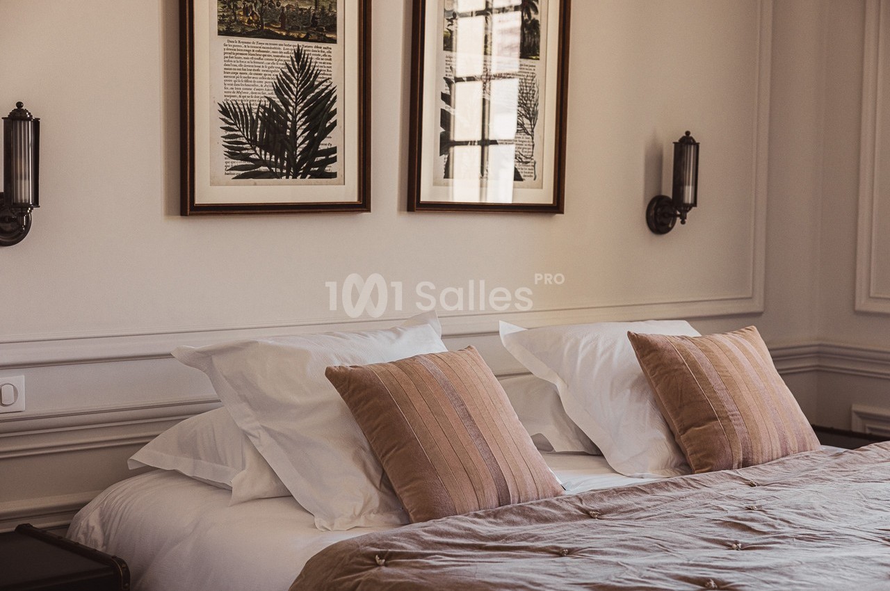 Lit double avec coussins beige et blancs, encadré par deux appliques murales et décoré de cadres au mur.