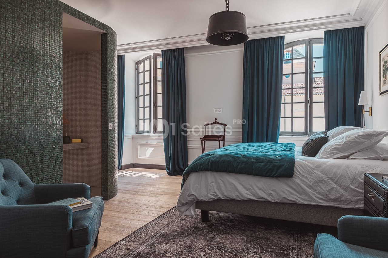 Chambre lumineuse avec lit double, fauteuils bleus, rideaux assortis et salle de bain attenante avec mosaïque verte.