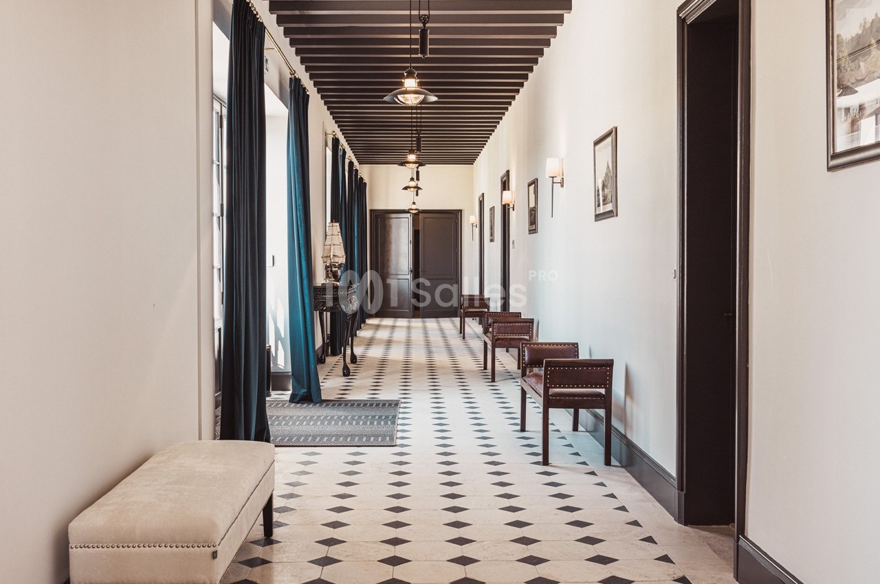 Couloir lumineux avec sol en carrelage noir et blanc, bancs en bois, rideaux sombres et éclairage suspendu.