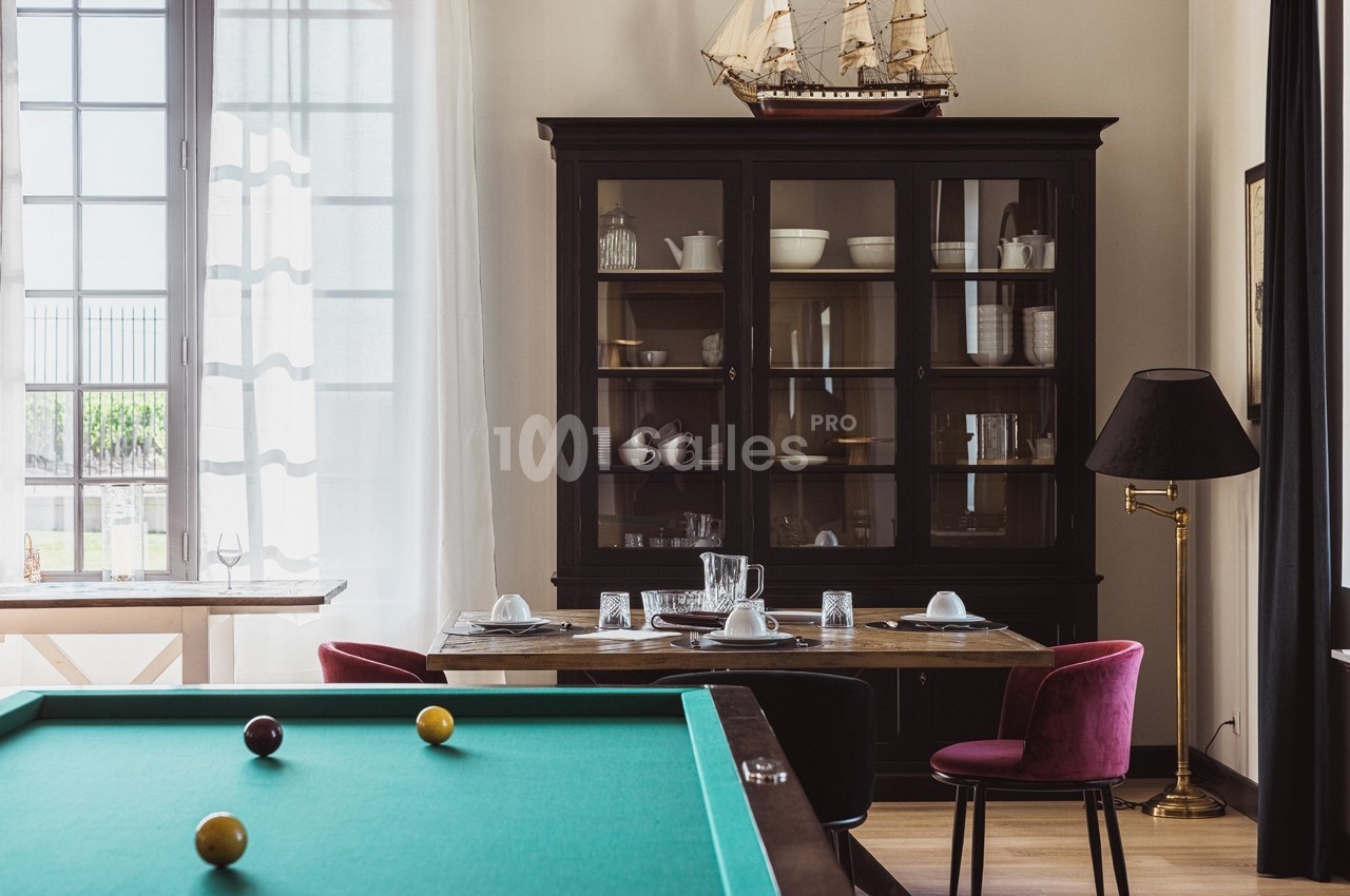 Salle à manger avec table dressée, vaisselier en bois sombre, billard vert au premier plan et voilure décorative.