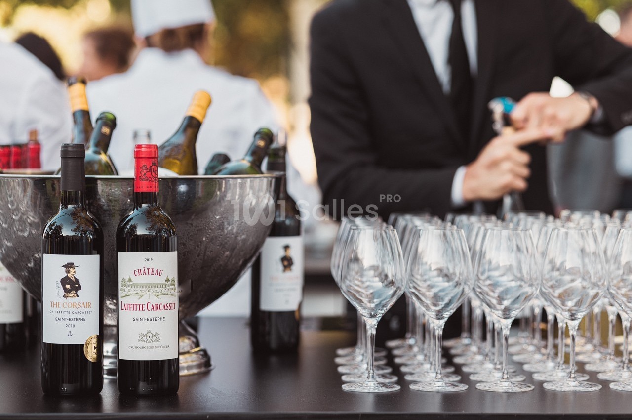 Bouteilles de vin rouge et seau à glace sur une table, avec des verres vides alignés et un serveur en arrière-plan.