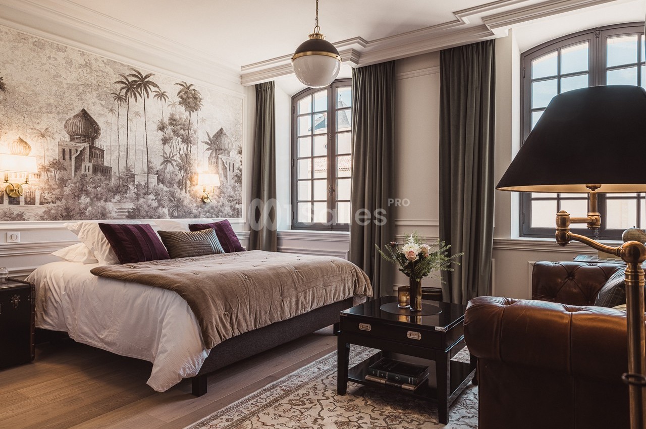Chambre élégante avec lit double, décoration murale exotique, grandes fenêtres, fauteuil en cuir et table basse fleurie.