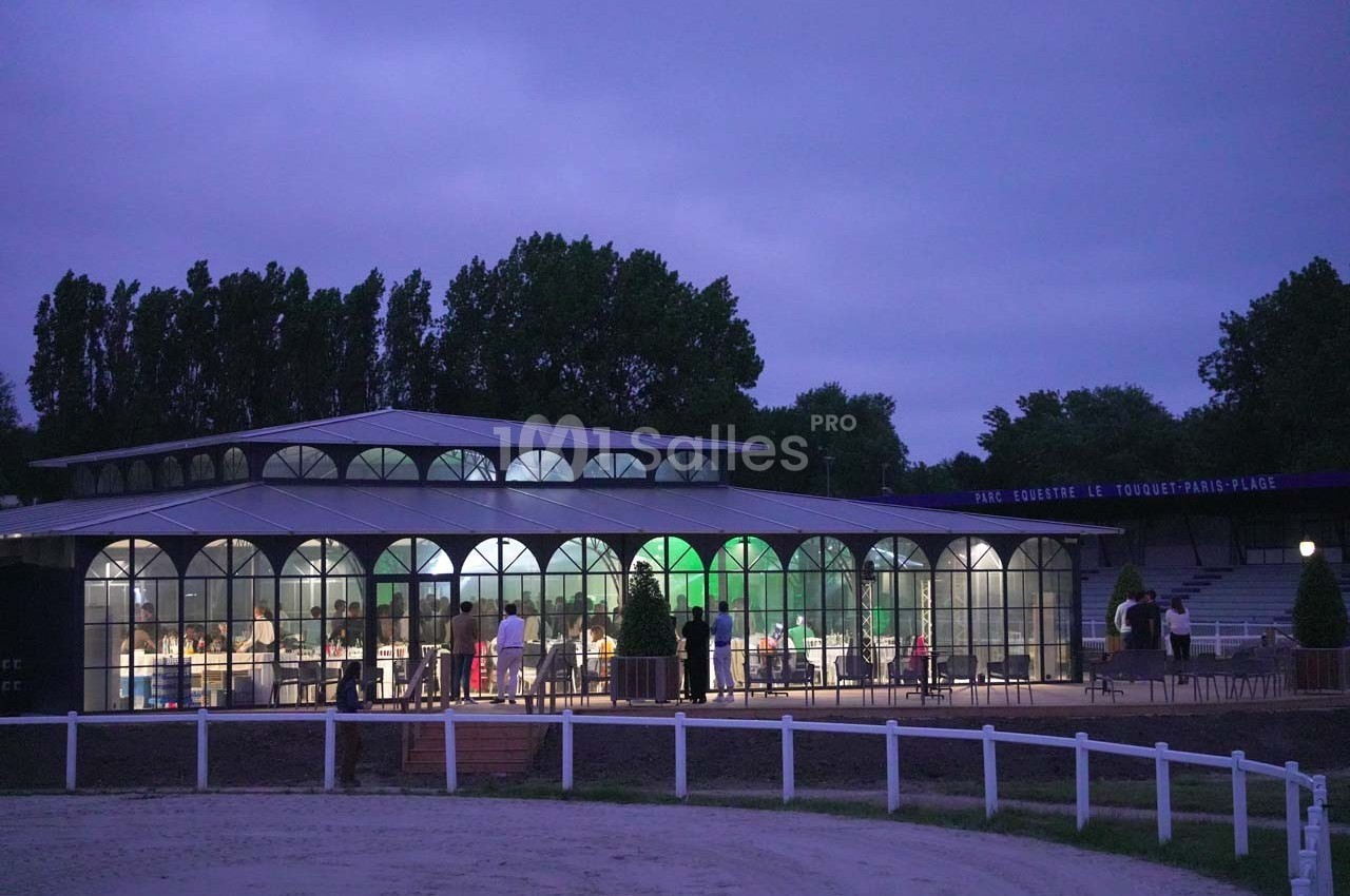 Bâtiment vitré éclairé en soirée, entouré d'arbres et d'une clôture blanche, avec quelques personnes à l'extérieur.