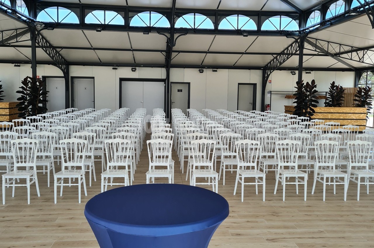 Salle aménagée avec des rangées de chaises blanches alignées devant une table recouverte d'une nappe bleue.