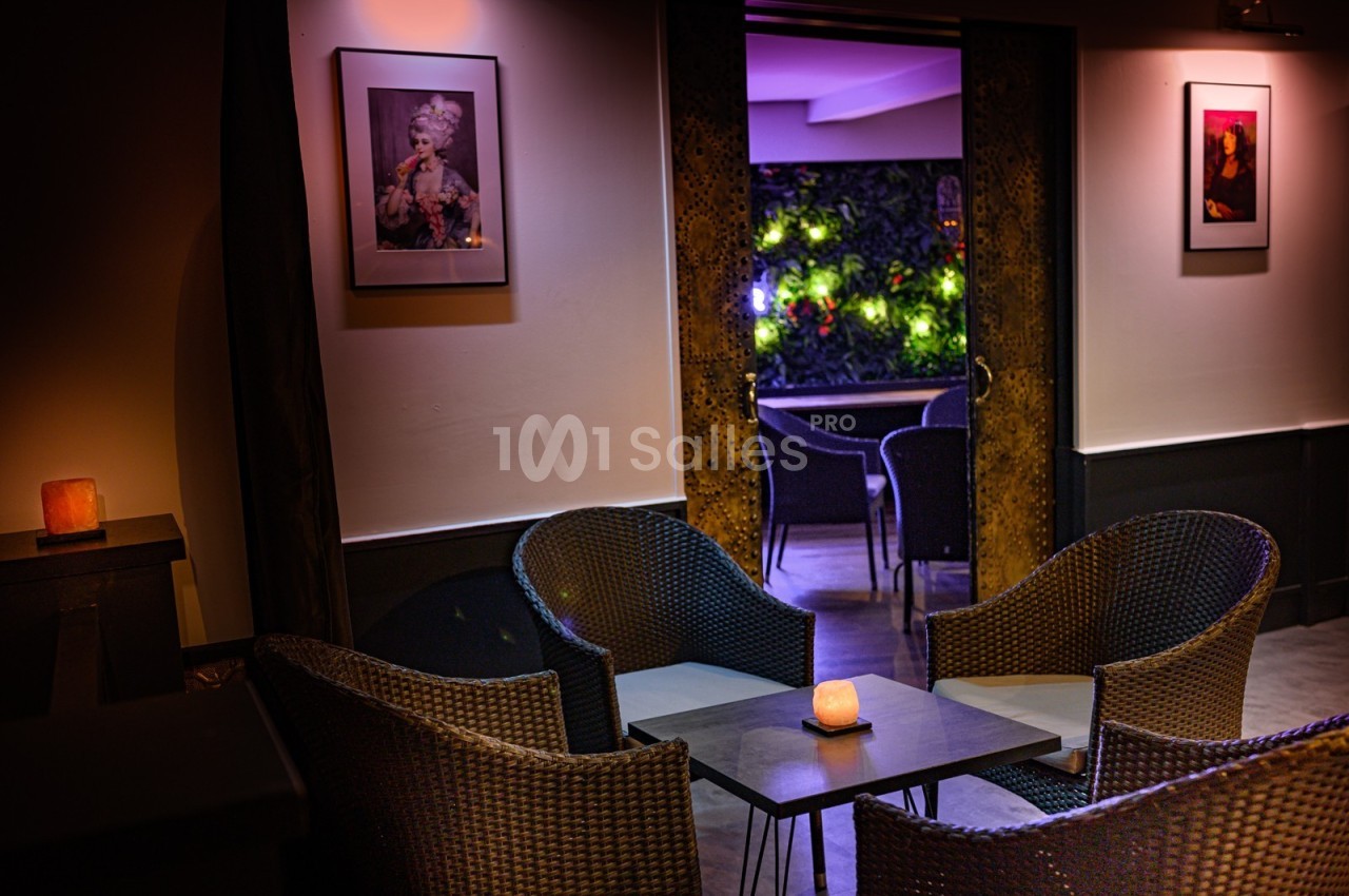Espace intérieur avec fauteuils en rotin autour d'une table, vue sur une terrasse éclairée et décorée de plantes.