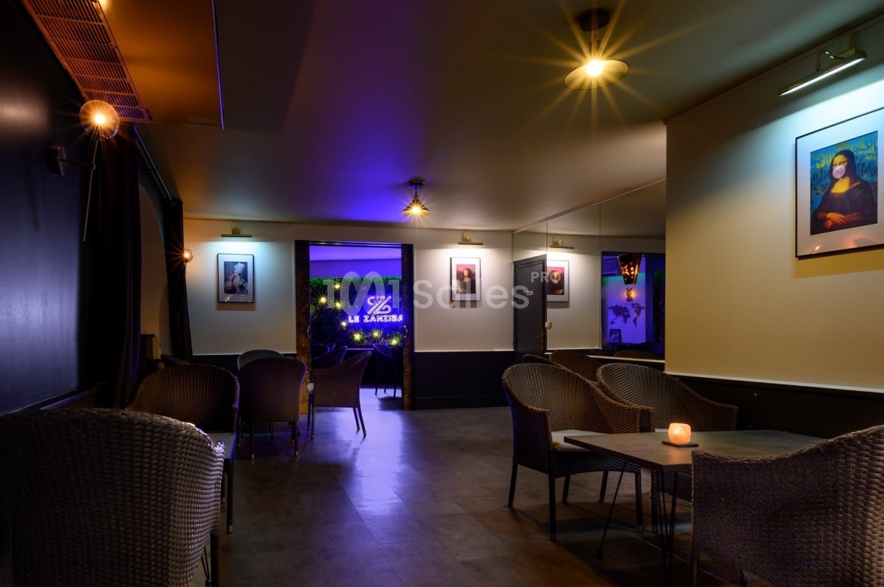 Salle de bar avec fauteuils en osier, éclairage tamisé et enseigne lumineuse visible au fond.