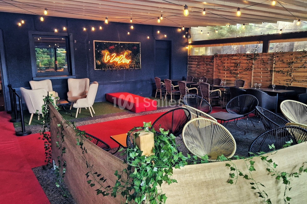 Espace extérieur aménagé avec fauteuils, tables, guirlandes lumineuses et panneau lumineux sur un mur sombre.