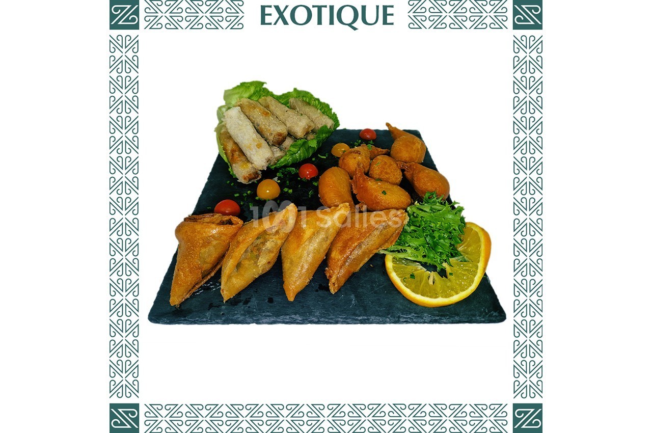Assortiment de bouchées salées comprenant samoussas, accras, nems, crudités et tranche d'orange sur ardoise.