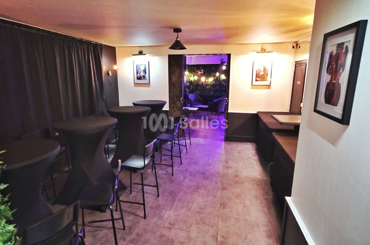 Salle avec tables hautes et tabourets, éclairage tamisé, décorations murales et rideaux noirs, ambiance intimiste.