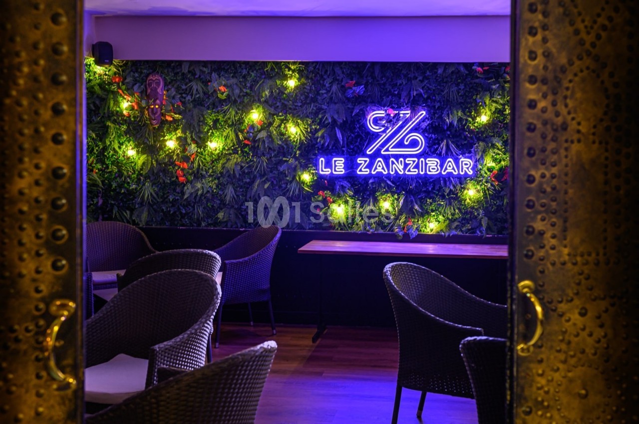 Mur végétal éclairé avec un néon ’Le Zanzibar’, entouré de fauteuils en osier dans un espace intérieur chaleureux.