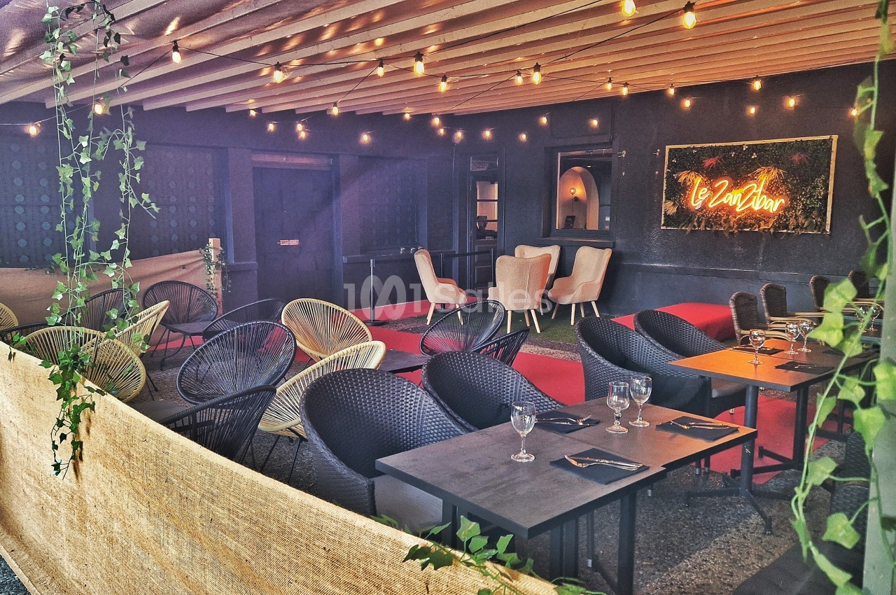 Terrasse couverte avec tables, chaises en rotin, guirlandes lumineuses et enseigne néon sur un mur sombre.