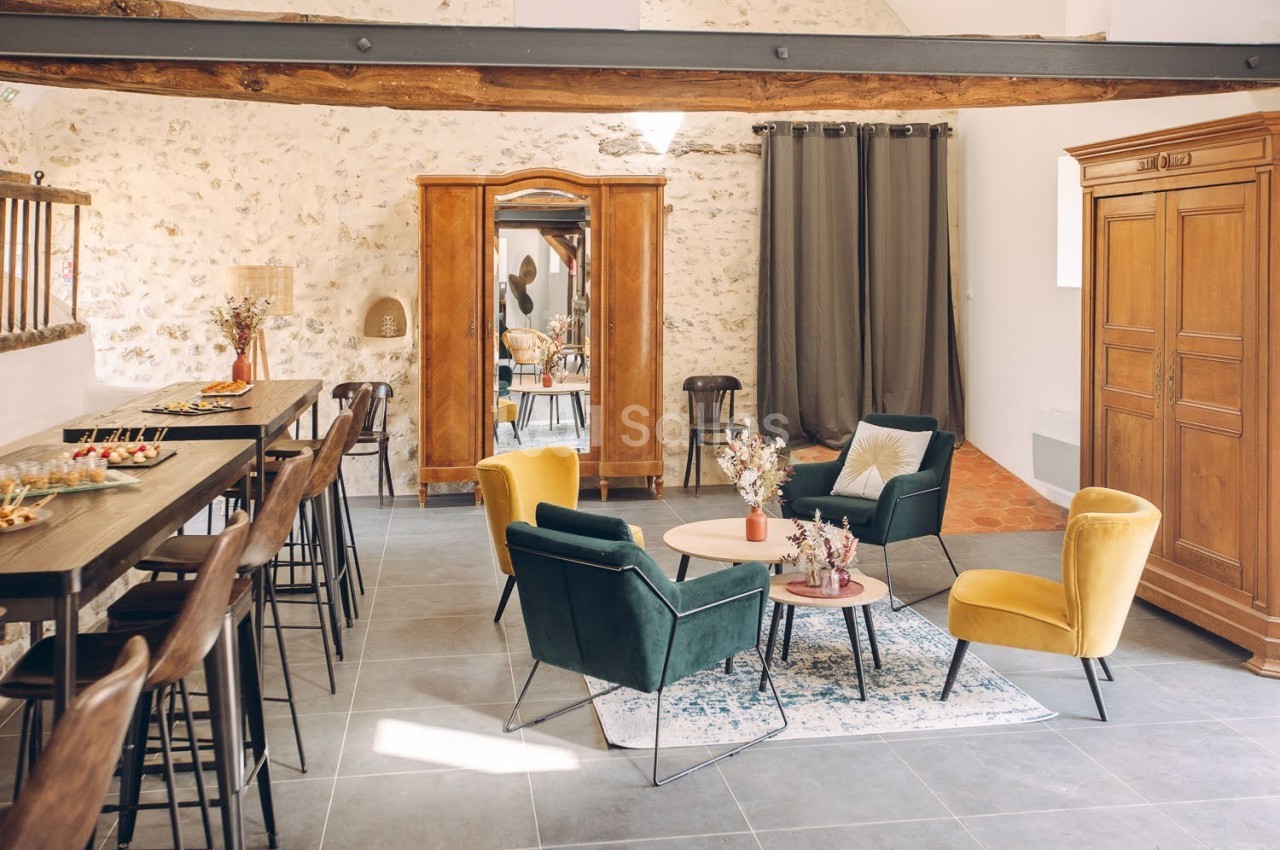 Salle lumineuse avec murs en pierre, mobilier moderne et ancien, table haute, fauteuils colorés et décorations florales.