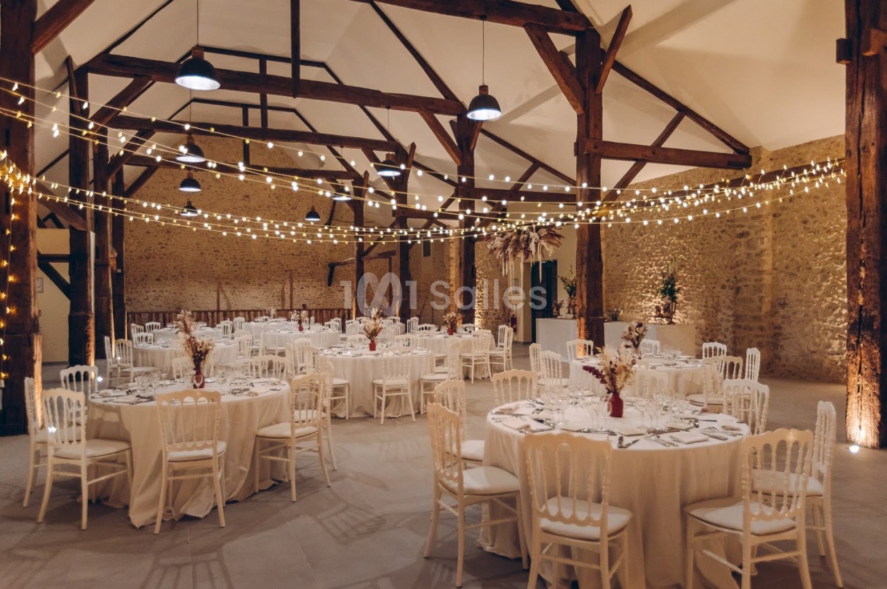 Salle de réception avec tables rondes dressées, chaises blanches et guirlandes lumineuses suspendues sous une charpente en…
