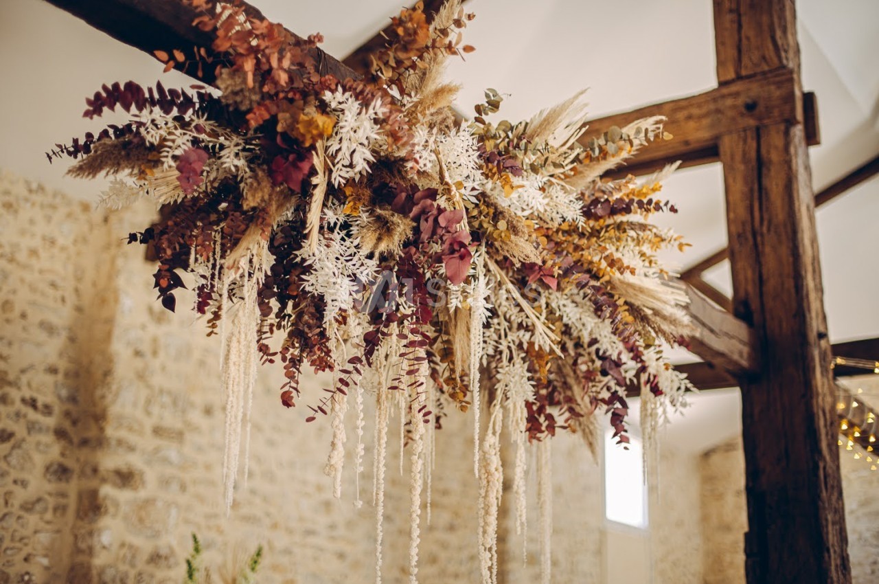 Suspension florale composée de feuilles séchées et de fleurs aux tons chauds, accrochée à une poutre en bois.