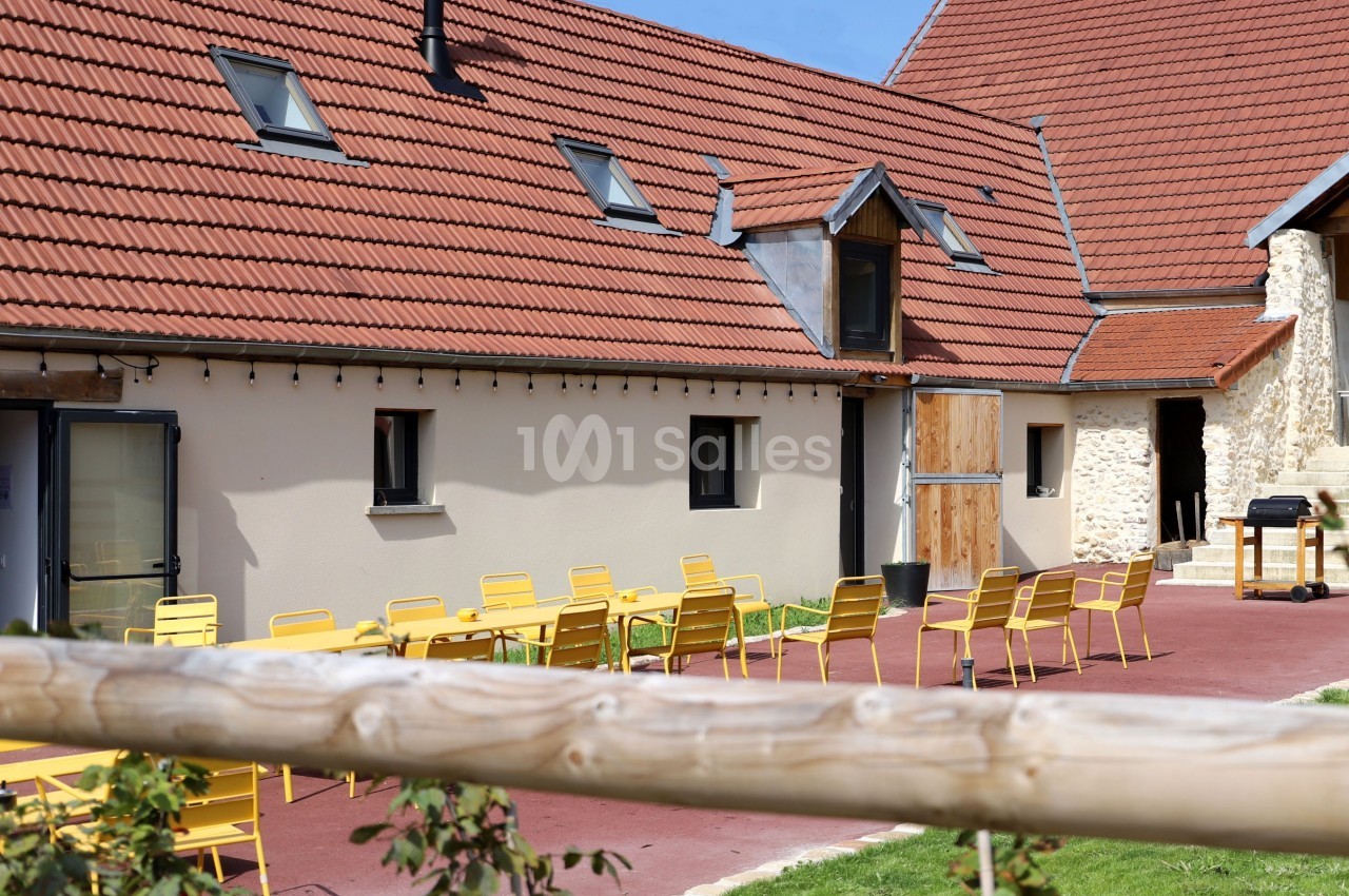 Cour extérieure avec des chaises jaunes disposées sur une terrasse en pavés rouges, entourée de bâtiments à toit en tuiles.