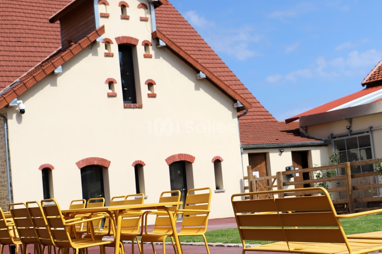 Façade d'un bâtiment à toit rouge avec une terrasse équipée de tables et chaises jaunes sur sol rouge.