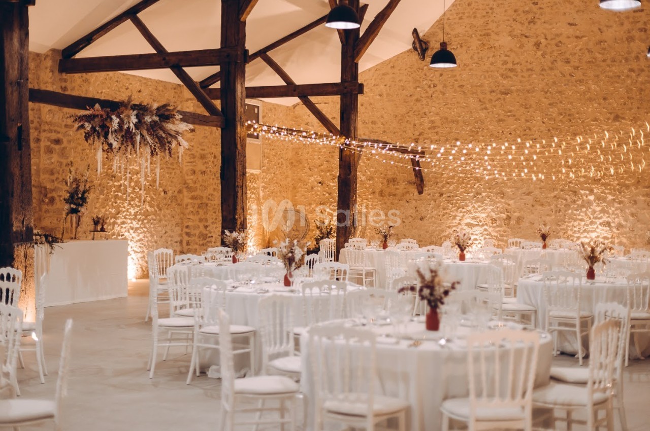 Salle de réception décorée avec des tables rondes, chaises blanches, guirlandes lumineuses et murs en pierre apparente.