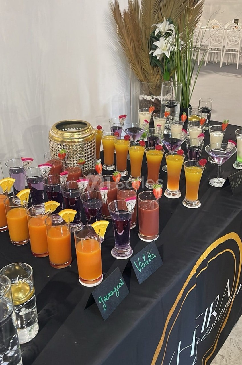 Table garnie de cocktails colorés avec décorations fruitées, étiquettes de saveurs et éléments décoratifs en arrière-plan.
