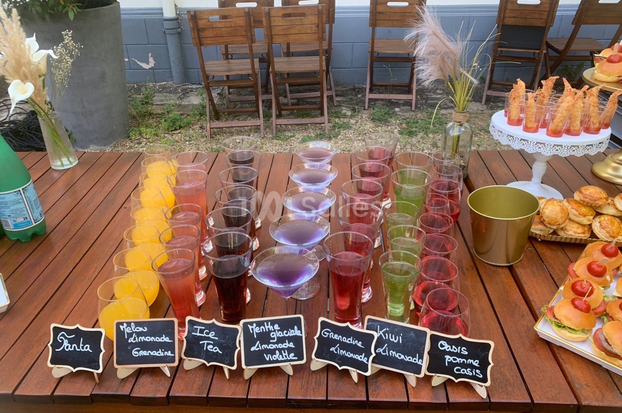Buffet de boissons colorées en gobelets alignés sur une table en bois, avec des étiquettes indiquant les saveurs.