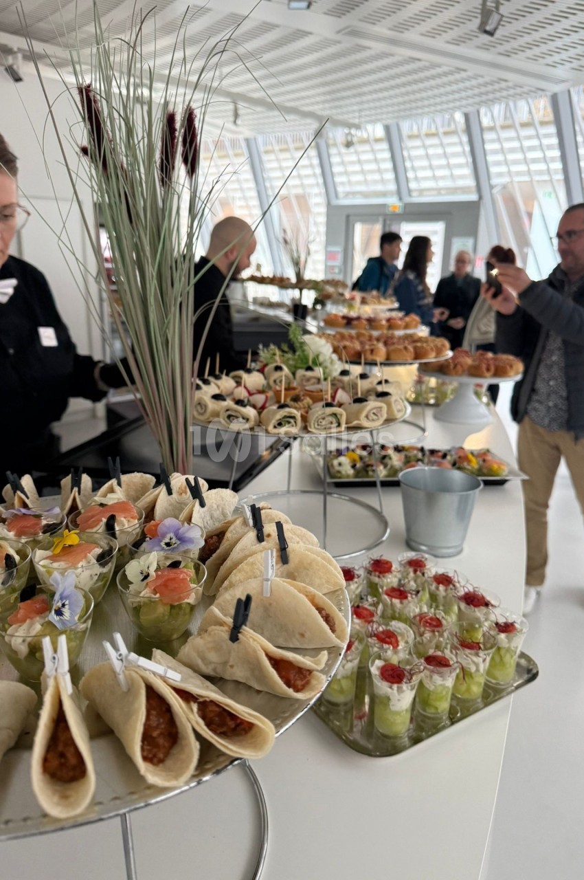 Buffet avec wraps, verrines et plats variés, présenté dans un espace lumineux avec des convives en arrière-plan.