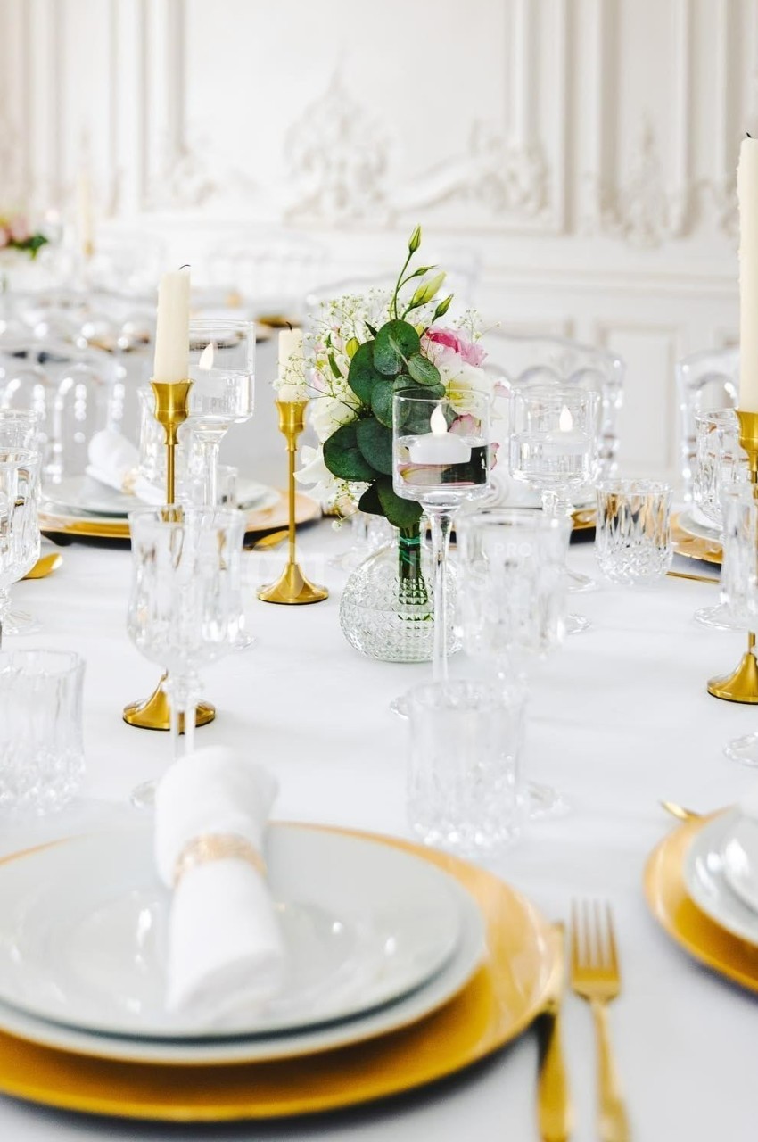 Table élégamment dressée avec vaisselle blanche, sous-assiettes dorées, chandeliers et bouquet floral central.