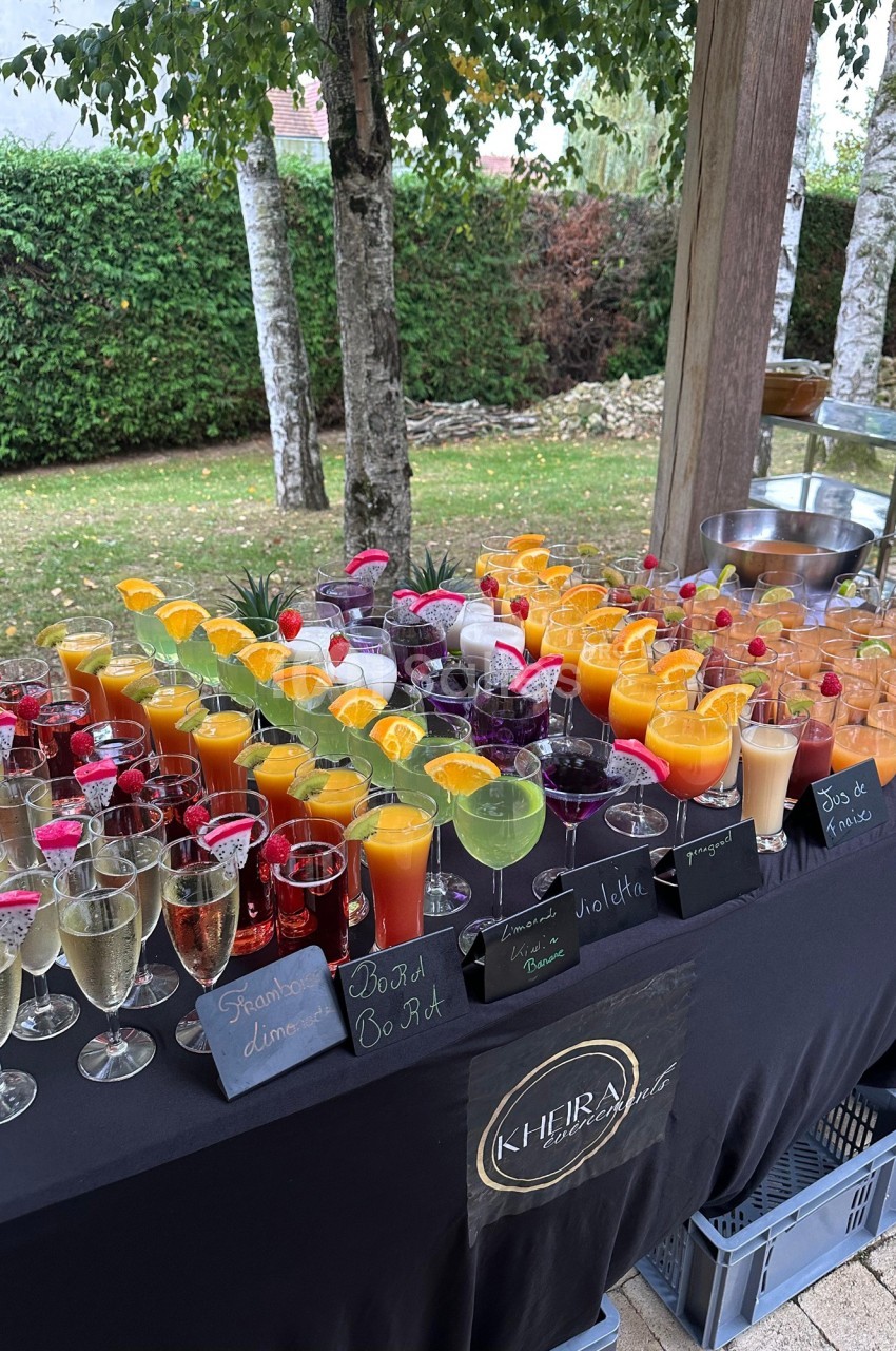 Table garnie de verres de cocktails colorés et de jus, disposés en extérieur près d'un jardin verdoyant.