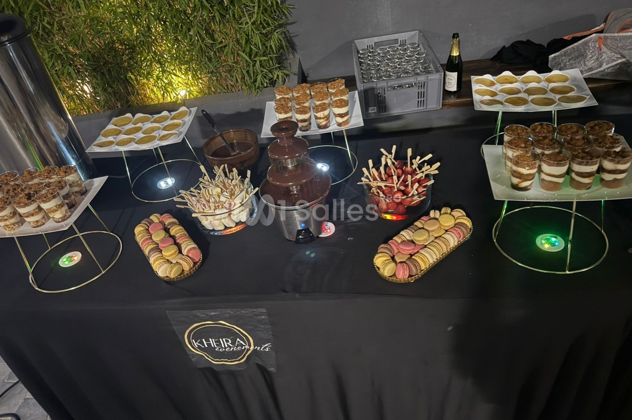 Table de desserts avec macarons, verrines, brochettes de fruits, fontaine de chocolat et boissons en arrière-plan.